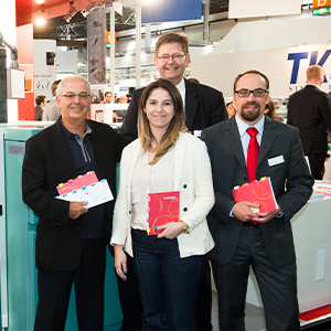 Na imagem, há a fotografia de quatro pessoas, sendo três homens e uma mulher. Todos usam roupas sociais e sorriem para a câmera, cada um com um caderno em suas mãos. Ao fundo, vê-se o cenário de uma feira em um centro de convenções.