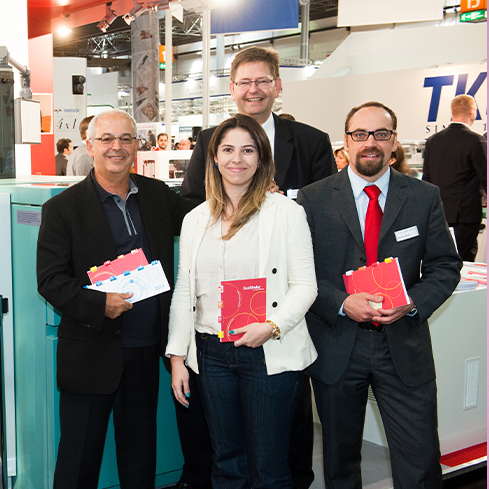 Na imagem, há a fotografia de quatro pessoas, sendo três homens e uma mulher. Todos usam roupas sociais e sorriem para a câmera, cada um com um caderno em suas mãos. Ao fundo, vê-se o cenário de uma feira em um centro de convenções.