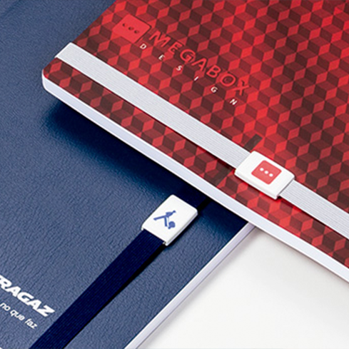 Na imagem, há a fotografia aproximada de dois cadernos executivos. A imagem foca nos detalhes das logomarcas de clientes da Ótima Gráfica, que aparecem em destaque como detalhes no elástico dos cadernos.